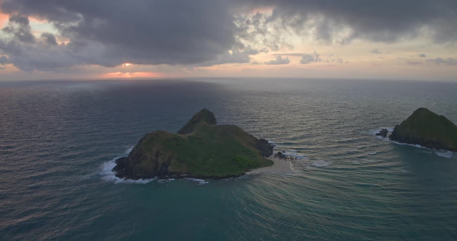 Slow aerial pullback reveals the iconic Mokulua Islands off the coast of Oahu, Hawaii, during a dramatic and cloudy sunrise