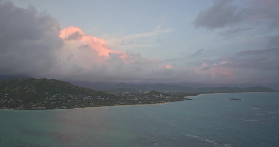 An aerial view of a tropical island coastline at sunset, with soft pink clouds hovering over the lush green mountains and turquoise sea
