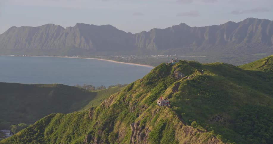 Epic aerial footage follows a hiking trail along a lush green mountain ridge, panning to reveal a stunning Hawaiian coastline