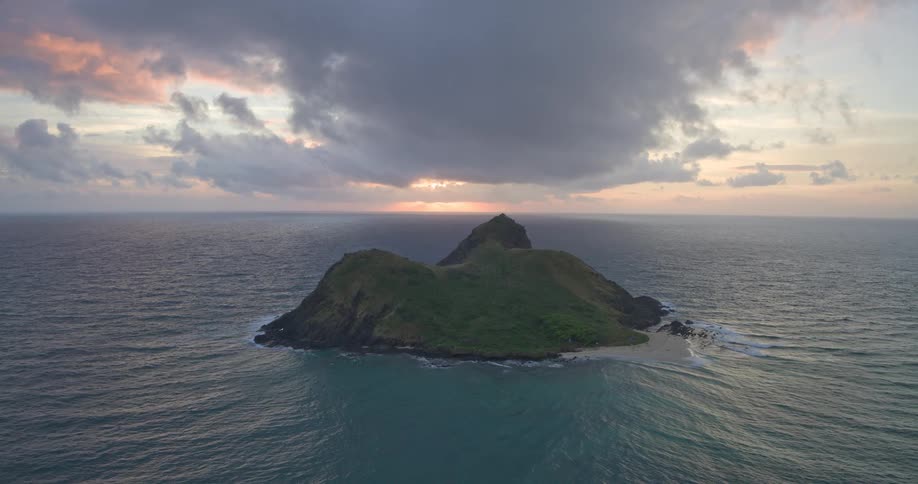 A serene aerial view shows a secluded tropical island in the vast ocean as the sun rises behind dramatic, moody clouds