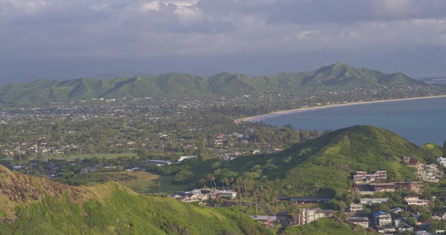 A stunning panoramic view from a high vantage point reveals a coastal town nestled between lush green mountains and the serene ocean in Hawaii