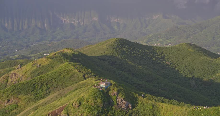 An epic aerial view pulls back from a group of hikers on a scenic green mountain ridge to reveal a vast, lush valley and a dramatic mountain range in the background