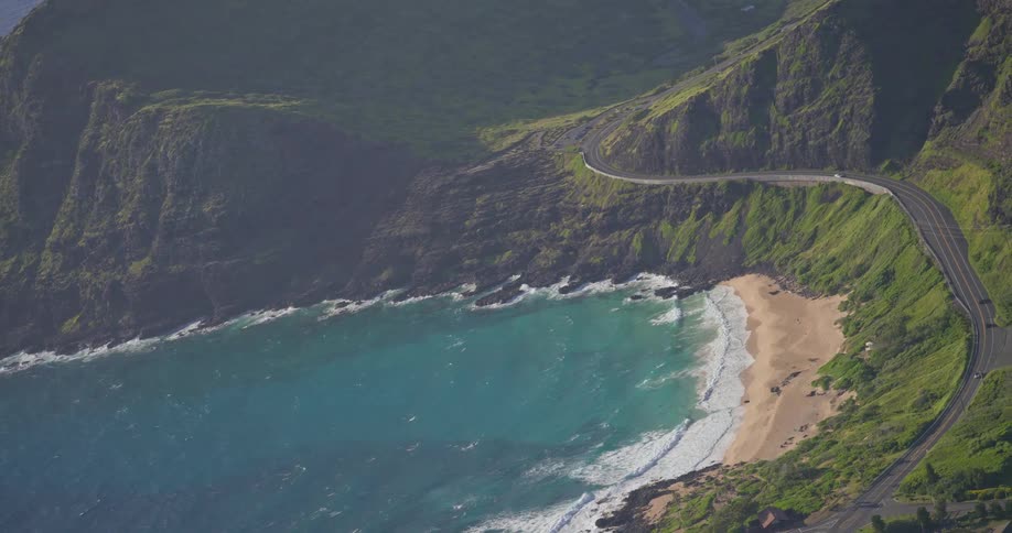An aerial view shows cars driving on a scenic highway along a lush, green coastline next to a secluded tropical beach