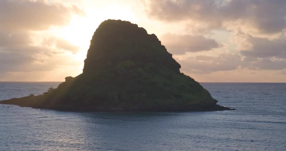 A beautiful time-lapse shows the sun rising from behind a silhouetted tropical island, casting brilliant sunbeams over the tranquil ocean