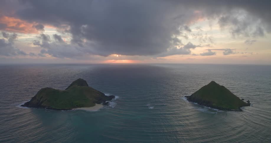 A majestic wide aerial shot of two tropical islands as the sun rises under a dramatic, cloudy sky over the calm ocean