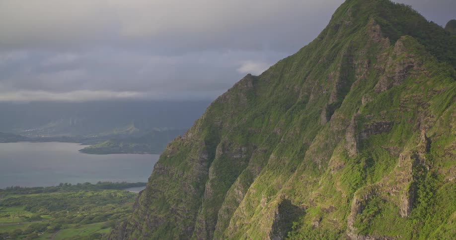 An epic vista of the dramatic, lush green cliffs of the Koolau Mountain Range on Oahu, with Kaneohe Bay in the distance under a cloudy sky