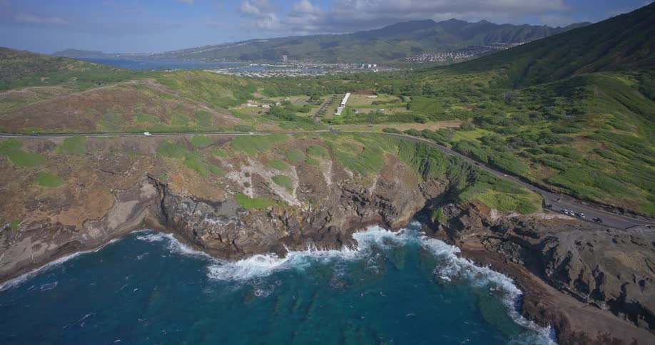 Wide aerial shot of the rugged Oahu coastline where turquoise ocean waves crash against the rocky cliffs below a scenic highway