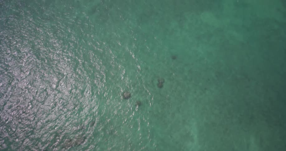 An aerial top-down shot of turquoise water slowly tilts up to reveal a stunning Hawaiian coastline with a beautiful beach and lush green mountains
