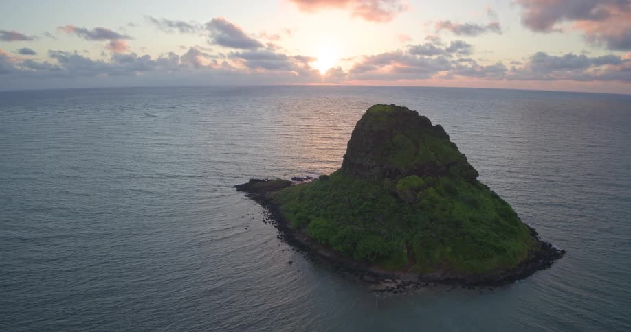 Aerial footage gracefully pulls back to reveal the iconic Mokoli'i island off the coast of Oahu, Hawaii, during a stunning sunrise over the ocean