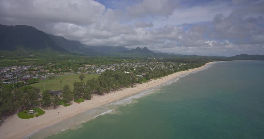 A sweeping aerial view of a pristine tropical coastline with a sandy beach, turquoise ocean, and lush green mountains under a partly cloudy sky