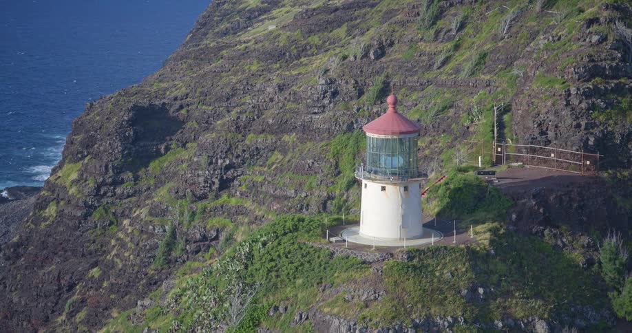 A steady telephoto shot zooms into the historic Makapuu Point Lighthouse perched on a rugged, green cliff overlooking the deep blue Pacific Ocean in Oahu, Hawaii
