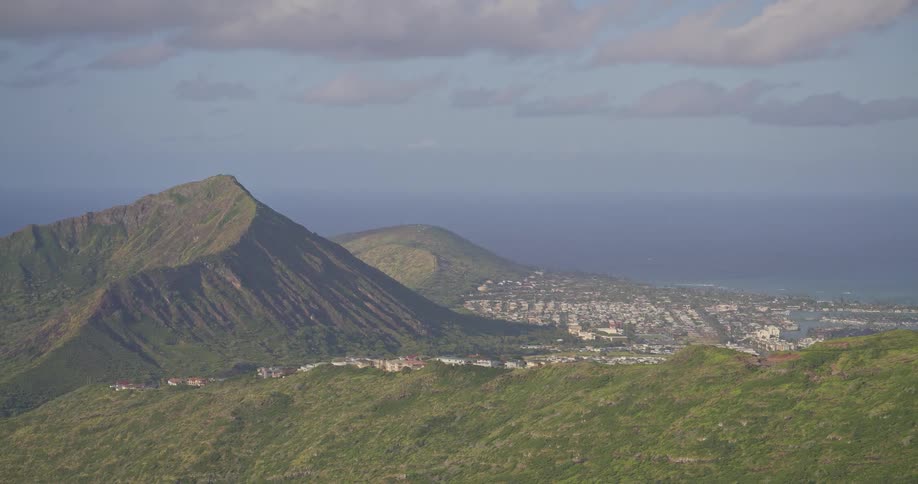 An expansive view from a mountain top shows a vibrant green Hawaiian landscape sloping down to a coastal town and the vast Pacific Ocean