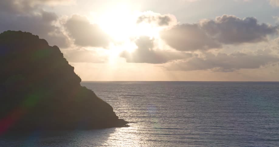 A serene sunrise sends golden rays through the clouds over a calm ocean, slowly revealing a silhouetted island