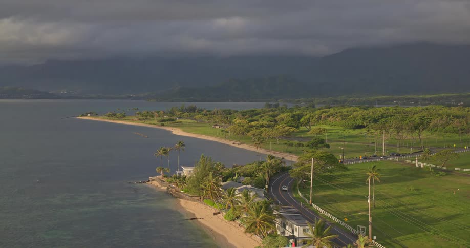 An aerial view showcases a scenic Hawaiian coastline with a road winding between the ocean and lush green fields, set against a backdrop of dramatic, cloud-covered mountains