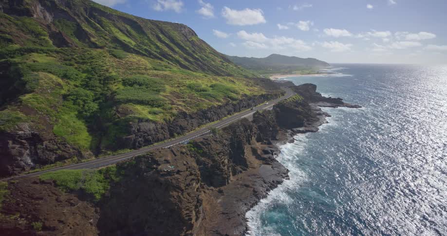 Epic aerial footage shows a scenic highway carved into the side of a volcanic, green mountain range overlooking the vast blue ocean