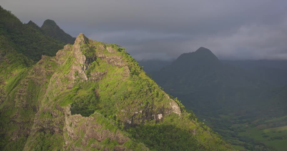 Aerial view pulls back from a dramatic, sunlit green mountain to reveal a vast valley under moody skies