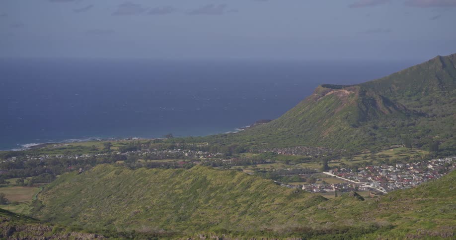 A slow zoom-out reveals the lush green mountains and a coastal town overlooking the vast blue ocean on a beautiful sunny day in Hawaii