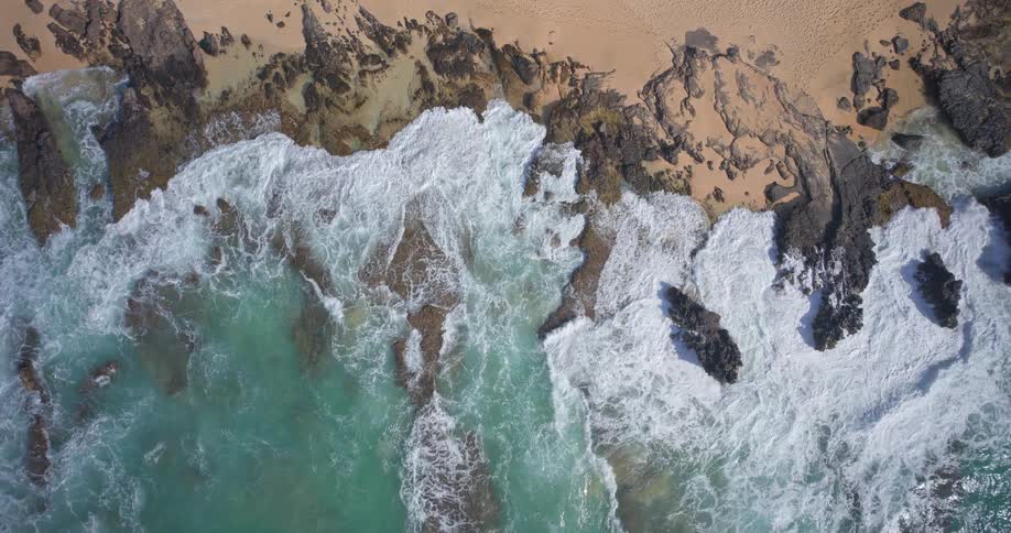 Ascending aerial top-down view of a dramatic rocky coastline where turquoise ocean waves crash against the shore
