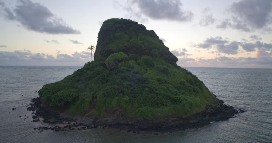 A slow aerial pull-back reveals the lush, green Mokoli'i island, also known as Chinaman's Hat, surrounded by the calm ocean off Oahu, Hawaii at sunrise