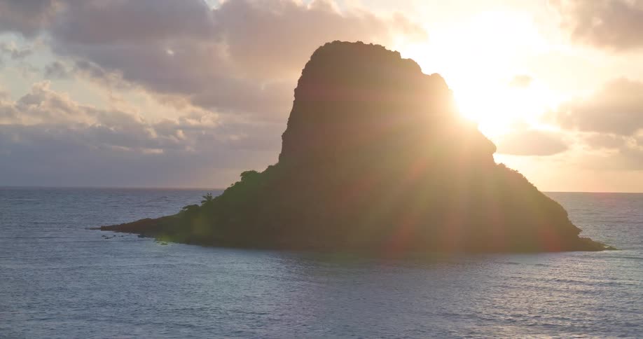 A slow aerial zoom reveals the silhouette of Mokoli'i island as the golden sun rises over the tranquil ocean in Hawaii