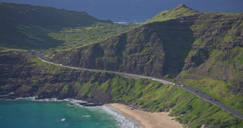 A scenic coastal highway winds along a lush green mountain, with cars driving beside the turquoise ocean and a secluded beach