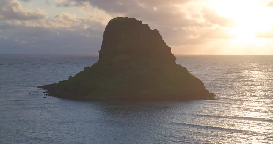 Aerial pull-back shot reveals a lush, tropical island silhouetted by the golden sunrise over the calm ocean