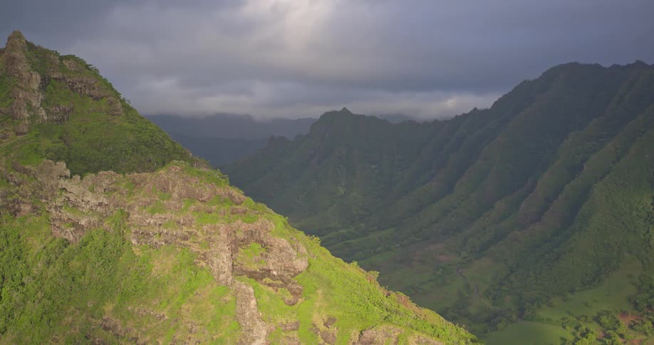 A dramatic aerial shot pans across a lush, sun-drenched mountain ridge, revealing a vast, moody valley under storm clouds