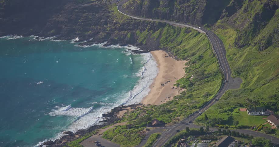 An aerial view captures the stunning beauty of a winding coastal highway alongside a secluded sandy beach with turquoise waves and lush green cliffs