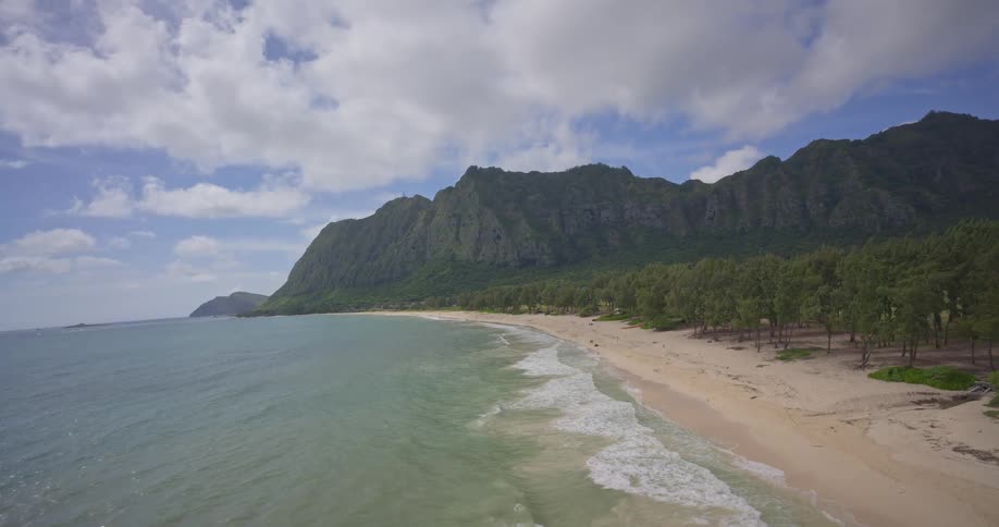 A stunning aerial view follows the coastline of a secluded tropical beach nestled against majestic green mountains on a beautiful day