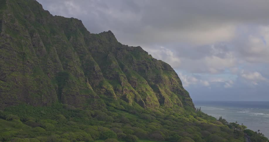 A breathtaking wide shot captures the majestic green mountains of Hawaii's coast meeting the ocean under a dramatic, cloudy sky