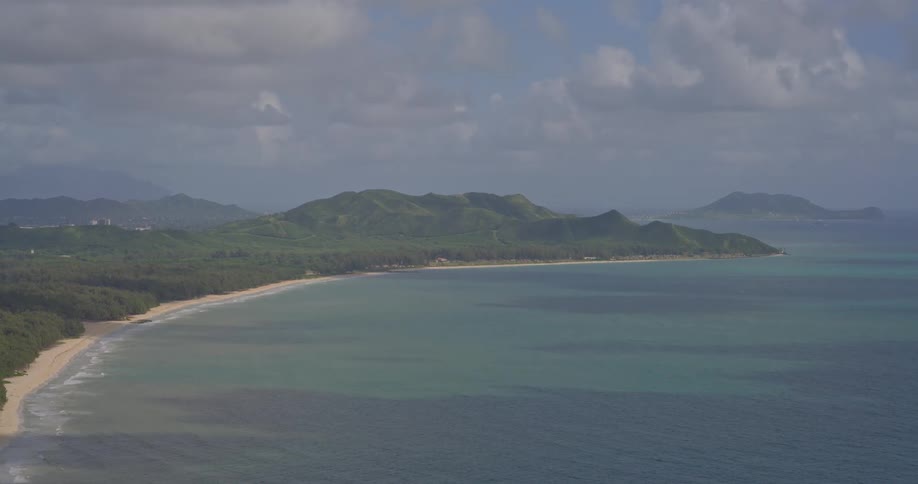 A scenic high-angle view of a tropical coastline, featuring a sandy beach, turquoise ocean, and lush green mountains under a cloudy sky