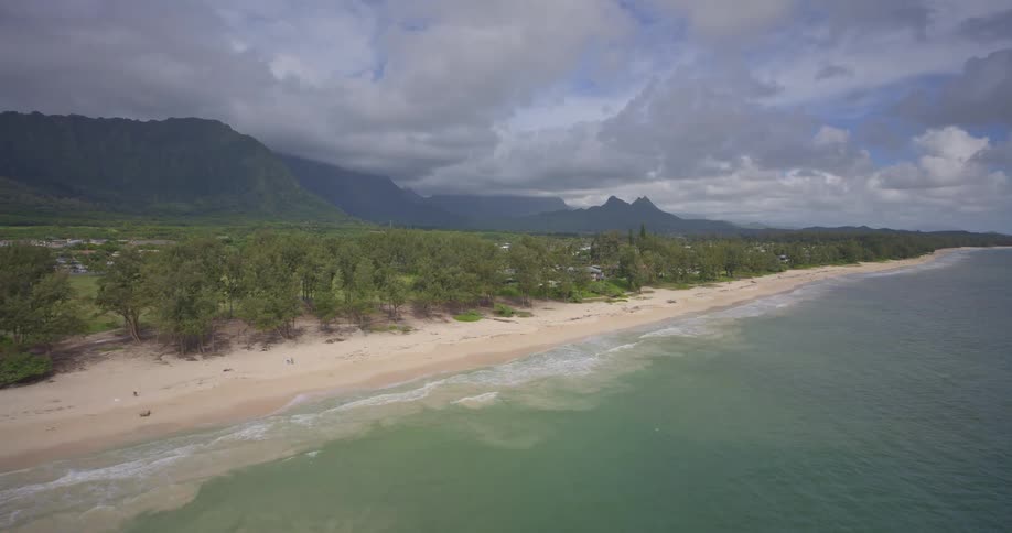 A beautiful aerial view flies along a sandy Hawaiian coastline with gentle waves, lush trees, and majestic green mountains in the background