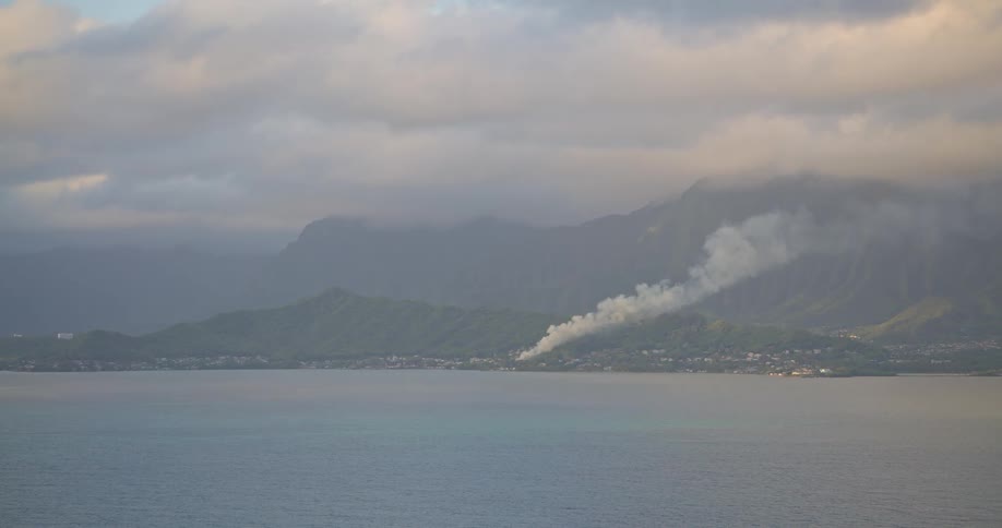 A plume of smoke from a distant fire rises over the green mountains of a tropical island coastline