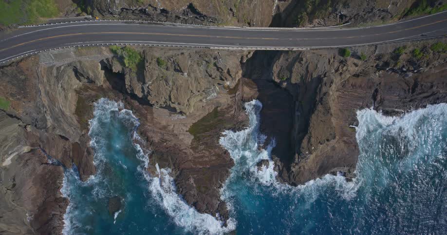 Aerial top-down view of a scenic coastal road running along a dramatic rocky cliffside with waves crashing below