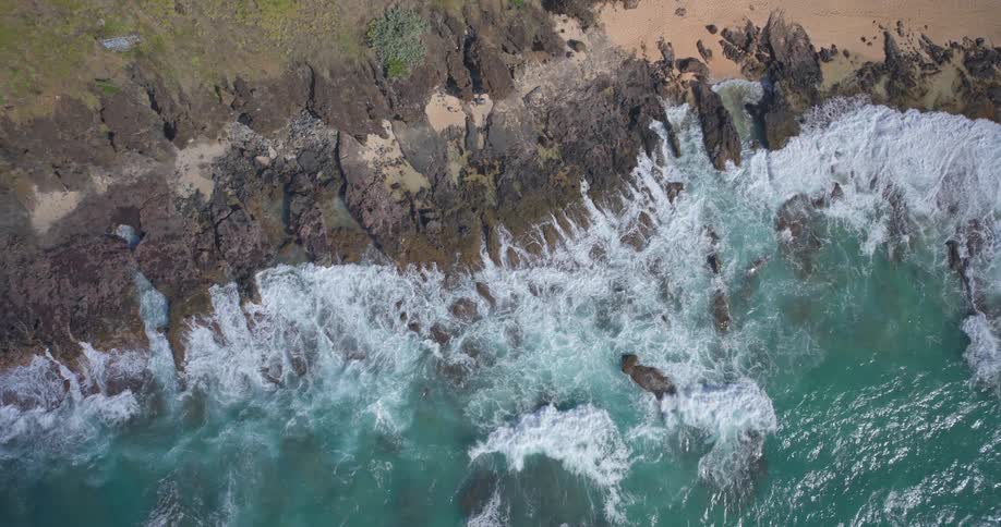 A stunning top-down aerial view of turquoise ocean waves crashing against a rugged rocky coastline and a sandy beach