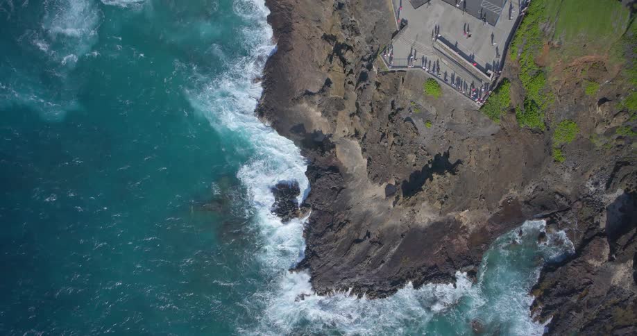 Top-down aerial view of powerful ocean waves crashing against a rocky coastline while tourists watch from a scenic overlook