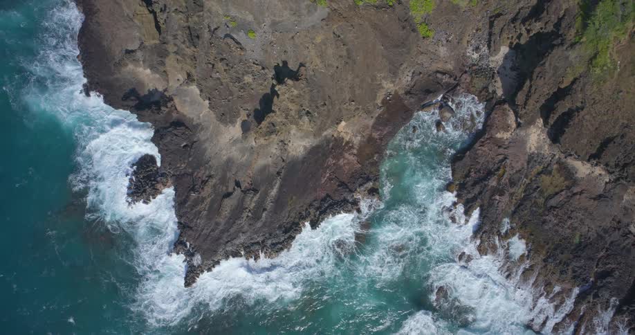 An aerial top-down view slowly ascends, revealing powerful turquoise ocean waves crashing against a rugged, rocky coastline and a tourist viewpoint