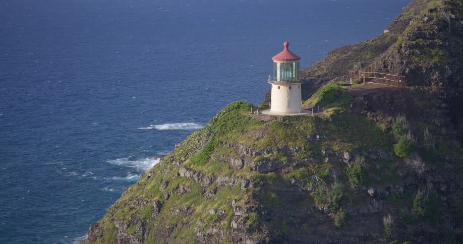 An aerial view slowly zooms out from the iconic Makapuu Point Lighthouse perched on a dramatic, lush cliff overlooking the Pacific Ocean in Hawaii