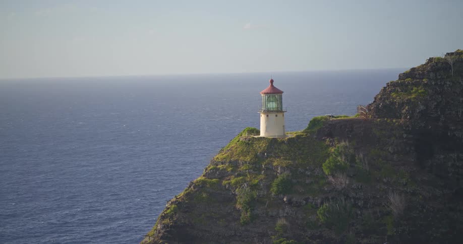 A lighthouse flashes its beacon from a grassy cliff overlooking the deep blue ocean as the camera levels its perspective