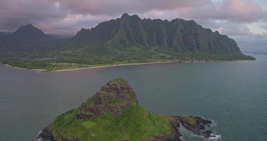 An aerial shot slowly pulls back to reveal the iconic Mokolii islet with the dramatic Koolau Mountains of Oahu, Hawaii in the background under a cloudy sky