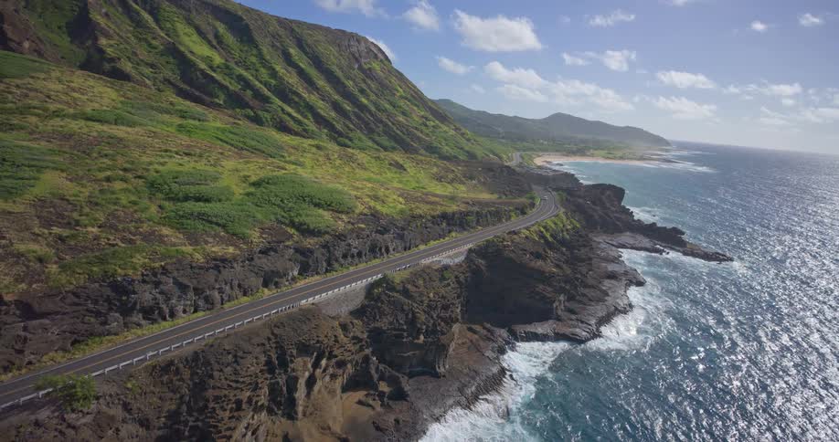 An epic aerial shot reveals a winding coastal highway along a lush, green mountain cliffside with the ocean waves crashing below