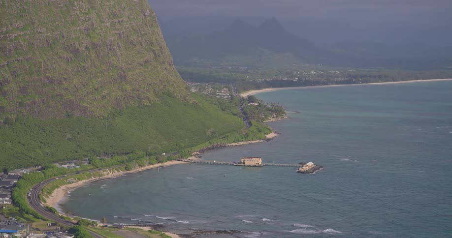 A breathtaking high-angle view captures the scenic coastline of Oahu, Hawaii, with a lush green mountain meeting the turquoise ocean and a pier extending into the bay