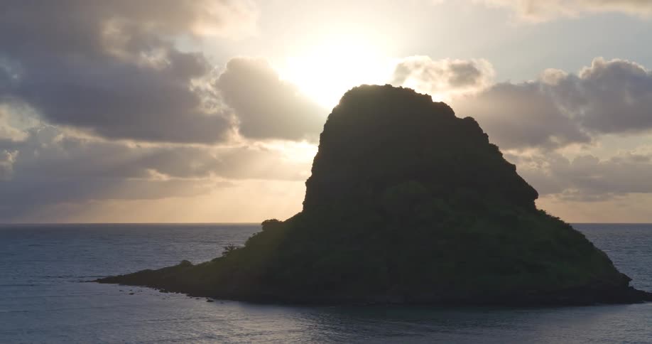 An aerial shot slowly pulls back from a silhouetted tropical island as the sun shines brightly through the clouds over the ocean