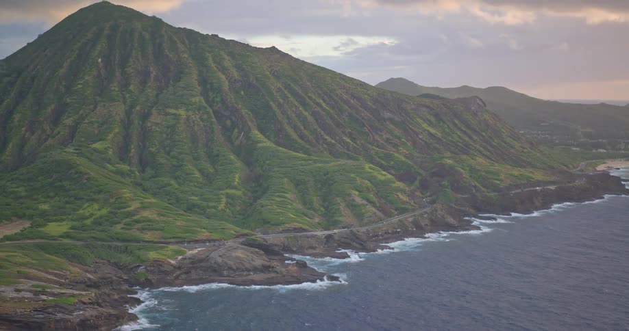 A breathtaking wide shot of a lush green mountain rising from the ocean along a scenic coastal highway in Hawaii at dusk