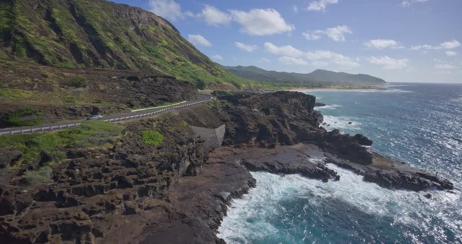 A picturesque aerial view of traffic on a coastal highway that winds along a rugged, volcanic shoreline next to a verdant mountain range