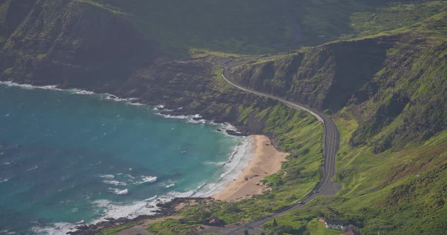 A scenic high-angle view shows cars driving on a winding coastal road alongside a beautiful Hawaiian beach with turquoise water
