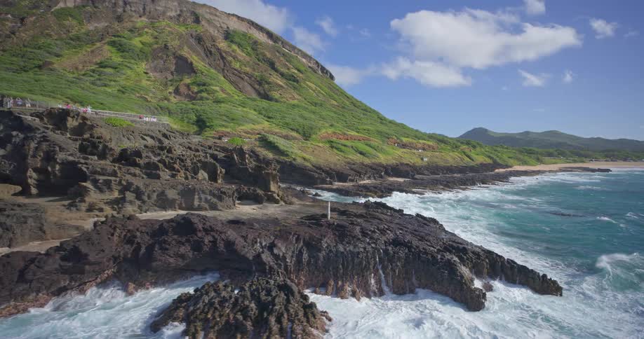 A stunning aerial view of the turquoise ocean crashing against the rocky shores of a lush green mountain in Hawaii
