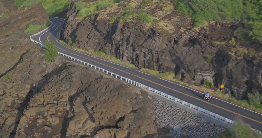 Drone follows a person on a moped driving along a breathtaking coastal road carved into rocky green hills by the ocean