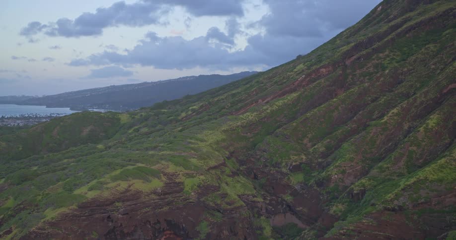 An aerial shot slowly pulls back over a vast, green mountain range to reveal the Hawaiian coastline and a city at dusk