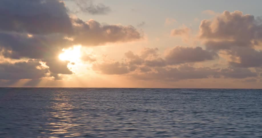 A breathtaking timelapse shows the sun rising over the calm ocean, breaking through the clouds with beautiful sunbeams
